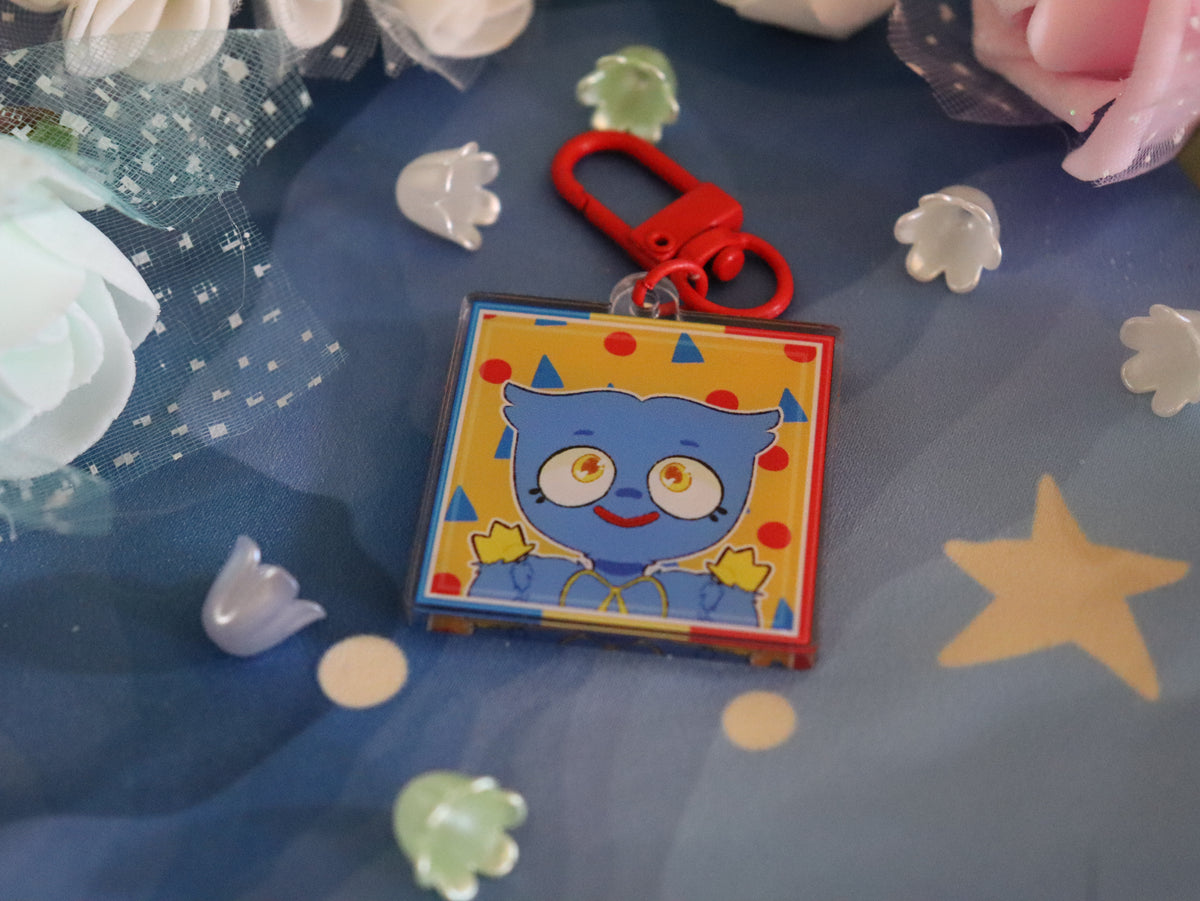 A square acrylic keychain charm featuring a blue cartoon monster character on a yellow background with red and blue accents, attached to a red metal clasp. The charm is displayed on a blue fabric background decorated with white and pink flowers and small translucent decorative pieces.
