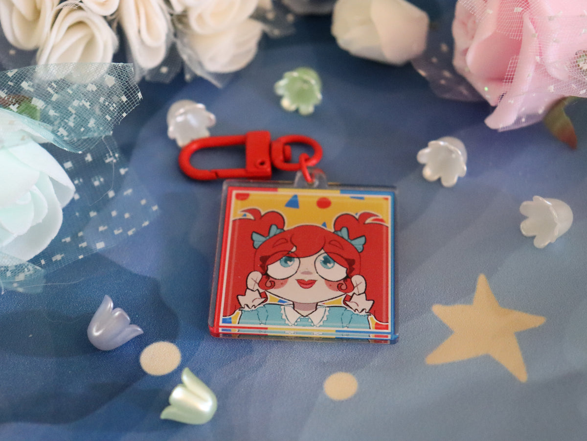 A square acrylic keychain featuring a colorful cartoon illustration of a girl with red hair and blue bows, attached to a red metal clasp. The keychain is displayed on a blue fabric background decorated with white and pink artificial flowers, small white flower petals, and a yellow star.